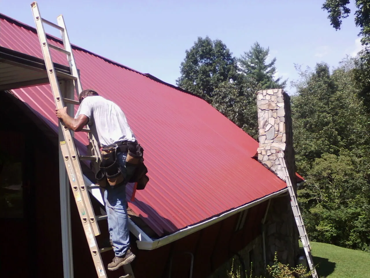 Roofer on a ladder beside a red metal roof and stone chimney — Soffit & Fascia Repair by Roofing Brothers in Staten Island